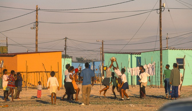 A group of people playing and gathering around with a soccer ball, next to orange and green houses.