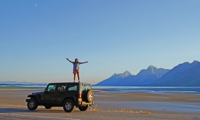 Grace DePaull standing on roof of jeep with arms outstretched, next to remote lake and mountains
