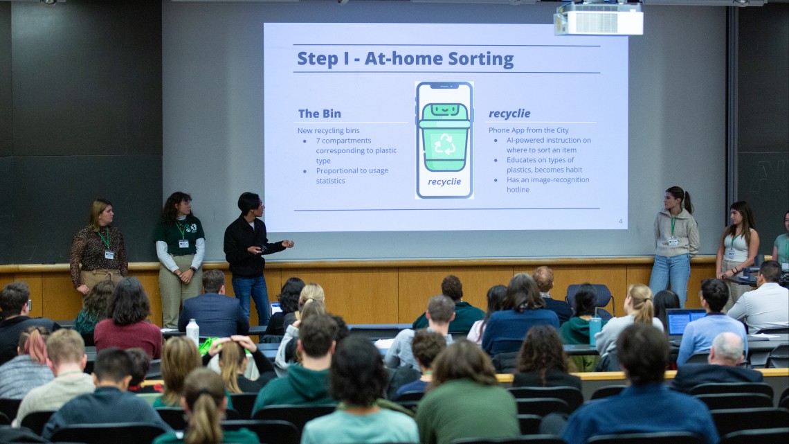 students present to an audience. A projection screen reads "Step 1: At-home sorting" with an illustration of a recyling bin