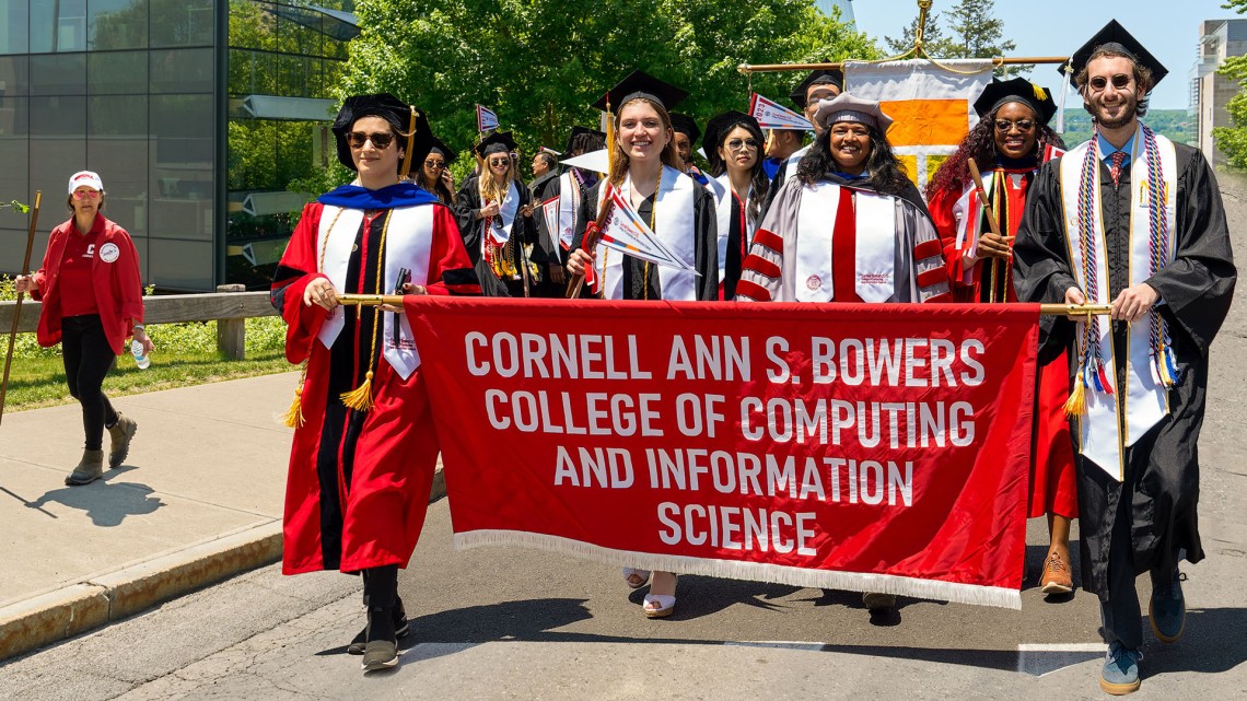 Several students in graduation robes stand behind a red banner on a sunny day.