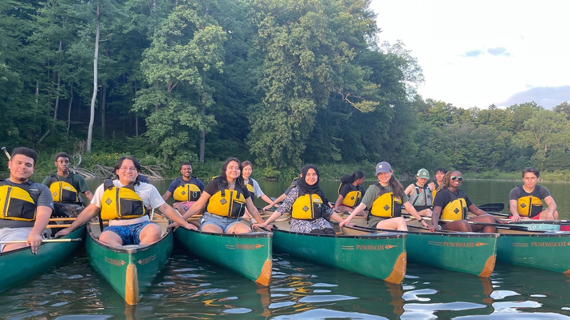 A dozen students sit in 6 green canoes on a lake.