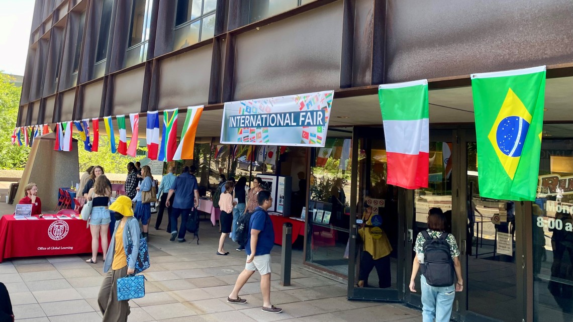 Students walk around Uris Hall terrace under global flags and an "International Fair" sign. 