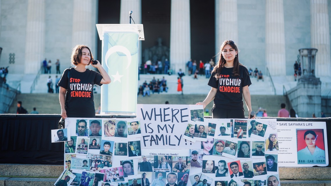 Two young people stand behind a "Where is my family?" sign and wear "Stop Uyghur Genocide" shirts in front of a large building with white columns.