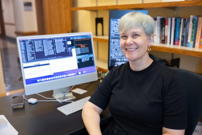 Person sitting at a desk with books behind and a large computer screen showing graphs