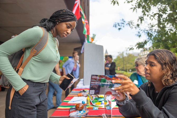 A student seated behind a table shows a postcard about the interntional relations minor to another student. 