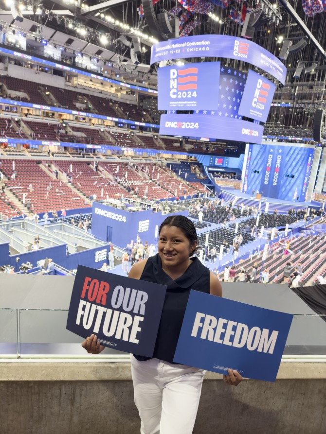 Student stands inside of the DNC Convention hall holding two signs that read "For Our Future" and "Freedom"