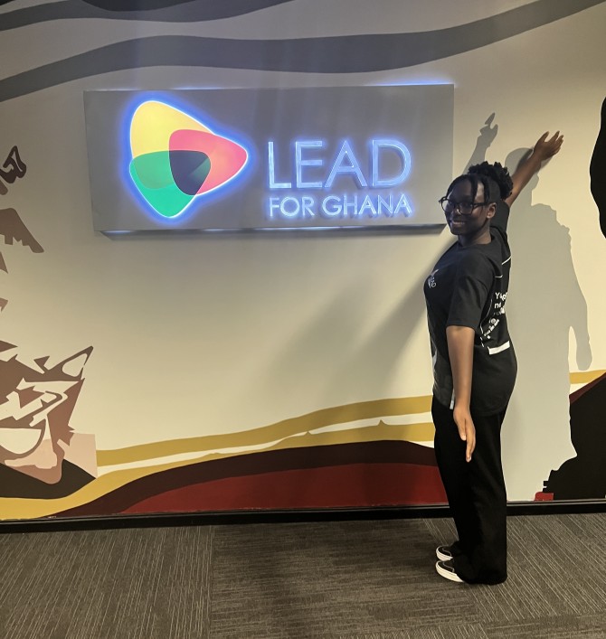 Eliana Amoh smiles with arms wide next to a "Lead for Ghana" sign.