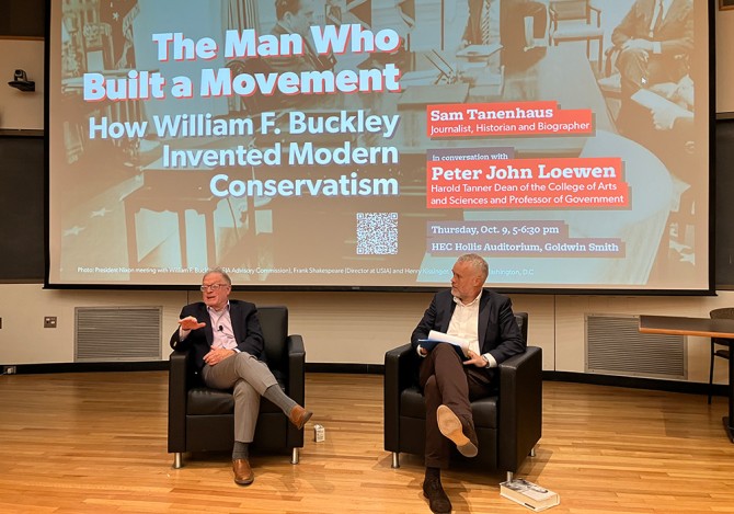 Two people sitting in arm chairs, conversing in front of an audience