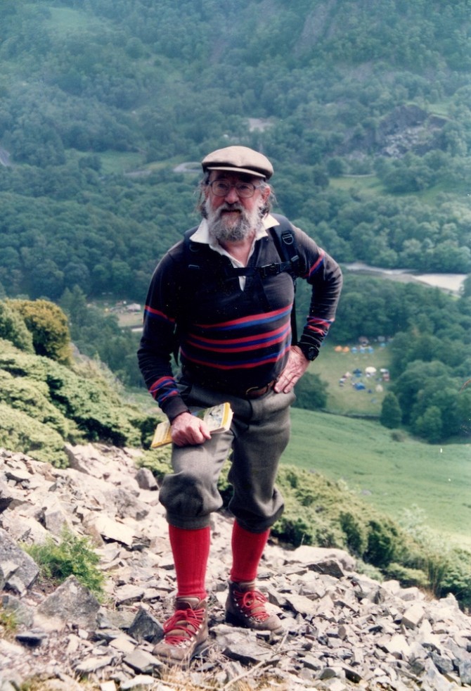 Person wearing a cap, sweater, wook hiking pants and sturdy boots standing on rocky ground high up on a very steep hill