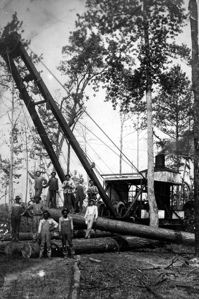 Black and white historical image from 1920 showing about ten lumber workers posing on large felled logs with a mechanical crane behind them