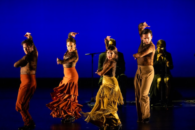 Four people dancing dramatically on a stage, wearing yellow and orange costumes