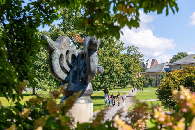 Large outdoor sculpture (an abstract shape somewhat like a butterfly) in the foreground with lawns, trees and pathways of a college campus quadtrangle in the distance. A few people walk on a path