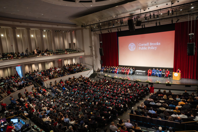 During the Brooks School graduation ceremony in Bailey Hall graduates, families, faculty, and guests gather in their seats to celebrate, a projector screen shows a logo with the text "Cornell Brooks Public Policy"