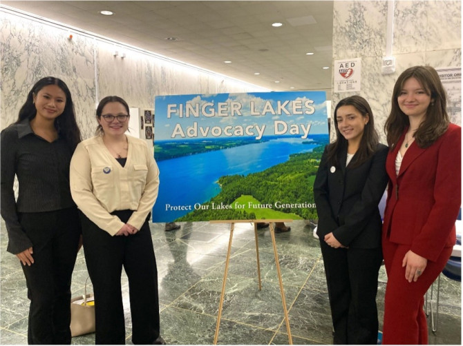 Left to right Isabelle Olaes, Pilar Seielstad, Madeleine Cierski, and Paige Peters standing alongside a poster for Finger Lakes Advocacy Day