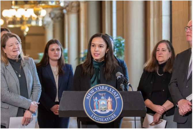 Sunni Horton  speaking delivering a speech at a NYS Legislature podium, with supporters standing around her
