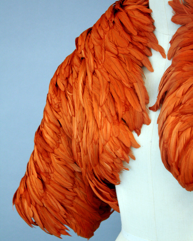 This orange feather bolero, with a short-waisted silhouette and three-quarter length sleeves, was shown as part of the 2007 Fall/Winter Ready-to-Wear collection. The surface is covered with overlapping rooster feathers arranged in diagonal bands radiating from the center back and neckline.