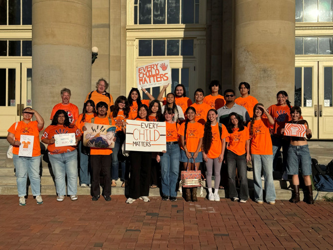 About 20 people stand on the stone steps of a building, wearing orange shirts and holding signs that say "every child matters"