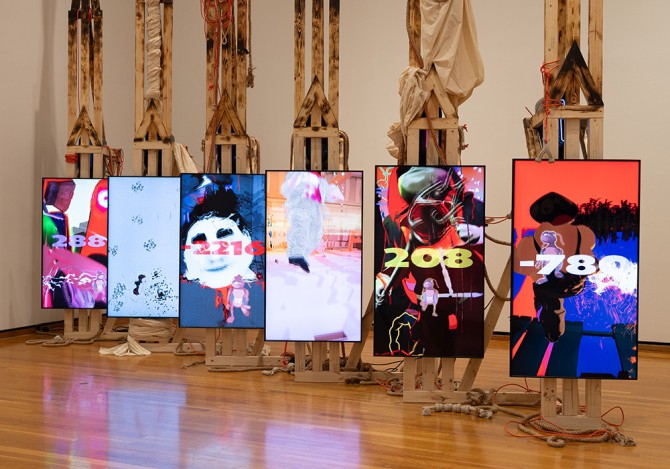 A large art installation featuring six vertically-oriented digital screens, each showing a different image in vivid colors