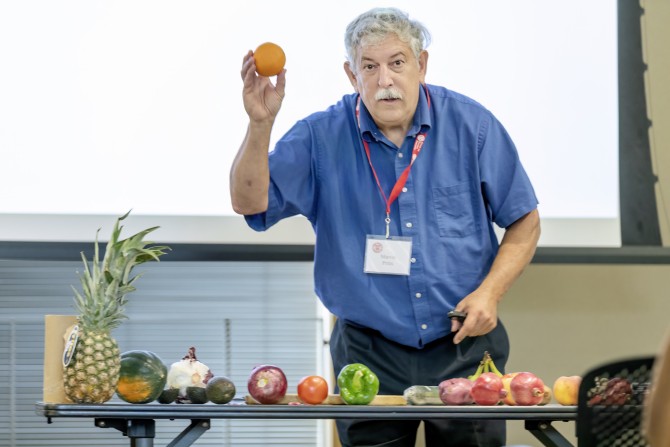 Professor Marvin Pritts stands in front of a row of fruit, holding up an orange.