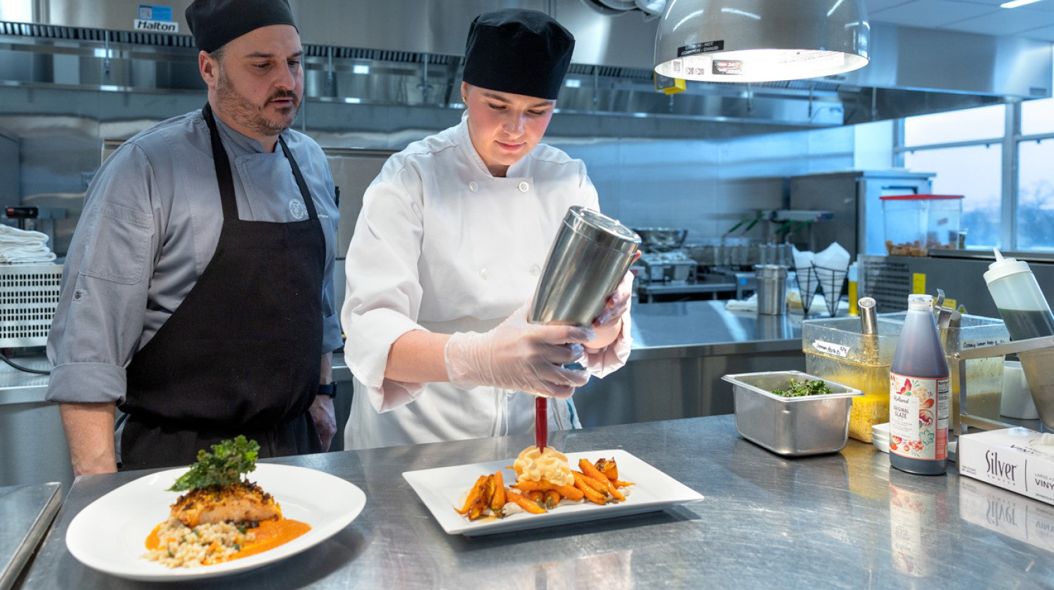 Chef instructor Christian Latimer (left) instructs Isabelle Louis ’26, a hotel administration major and chef assistant, how to add the finishing touch to the roasted carrots appetizer.
