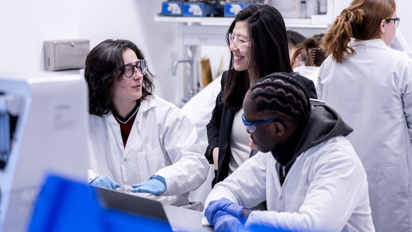A smiling professor consults with two students in a lab environment