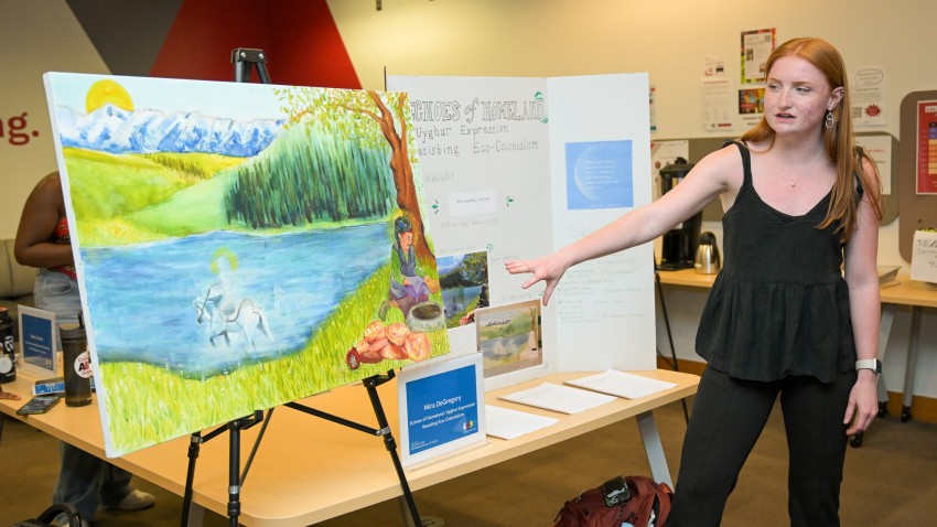 Student Mira DeGregory gestures toward a colorful painting of a person sitting in grass near a lake. 