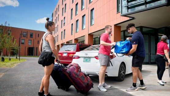 Three people carrying items and pulling luggage outside a residence hall. 