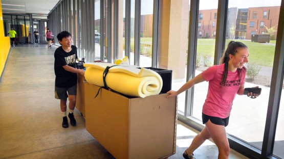 A young woman pulls a moving bin through a hallway with a young man pushing from behind. 