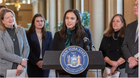 Sunni Horton speaking delivering a speech at a NYS Legislature podium, with supporters standing around her