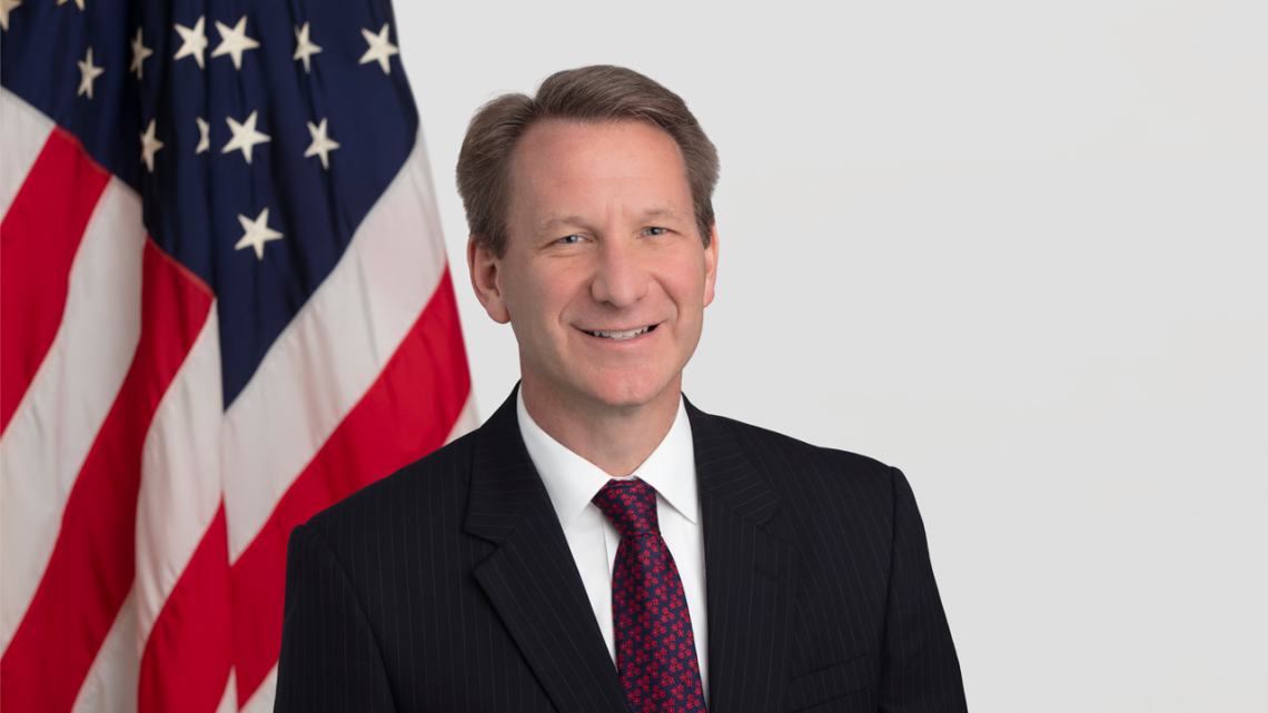 A headshot of Dr. Norman Sharpless, who is wearing a suit and tie, and seated in front of the American flag