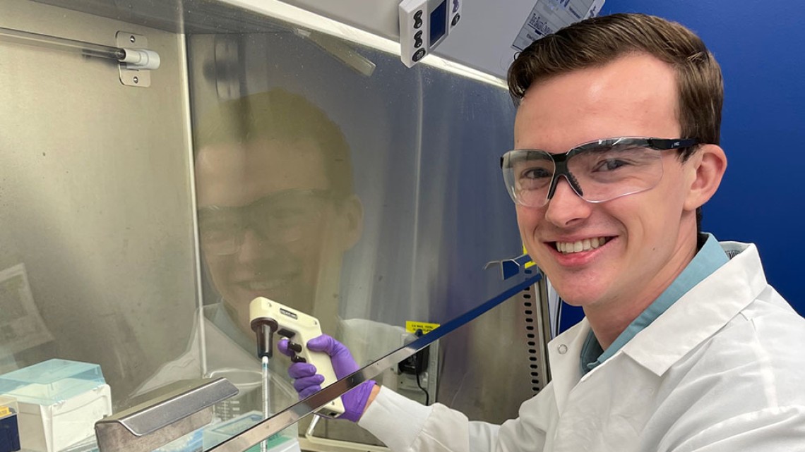 Jackson Bauer holds a tool in a lab setting while wearing a white lab coat and glasses. 