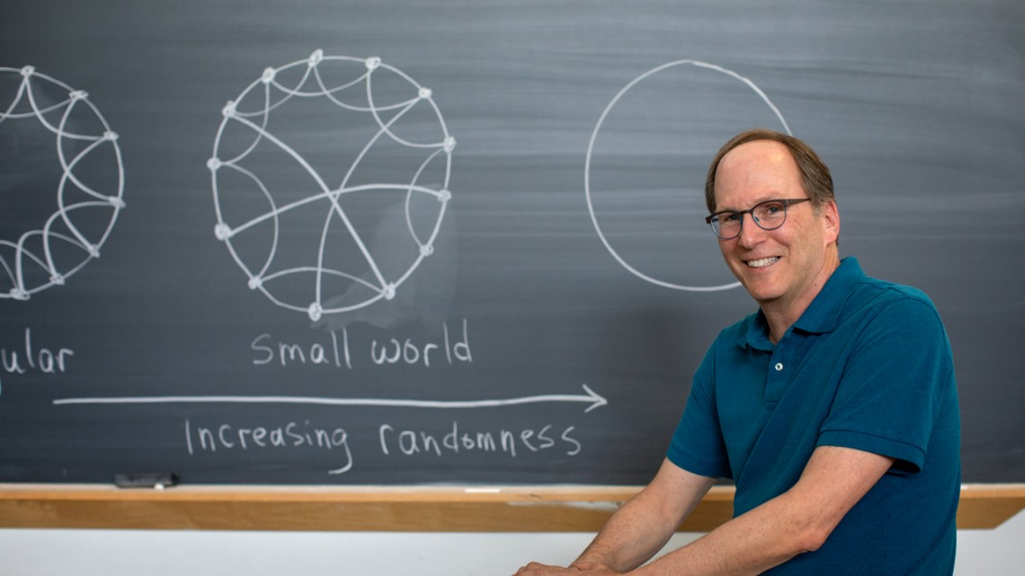 Steve Strogatz sitting in front of blackboard with small world diagram