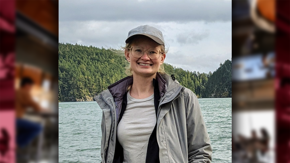 The head and torso of a blond woman in a grey coat and ball cap in front of a lake