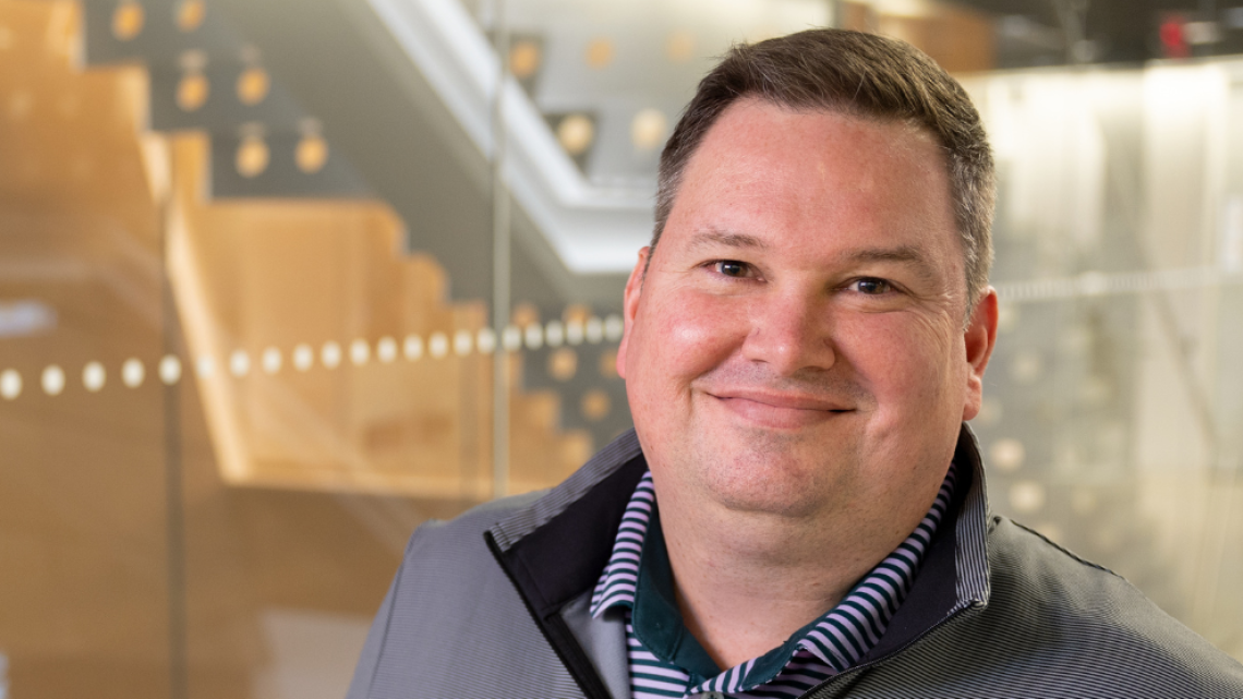 Associate Chief Information Security Officer Tim Bradish standing in hallway of academic building, smiling for the camera.