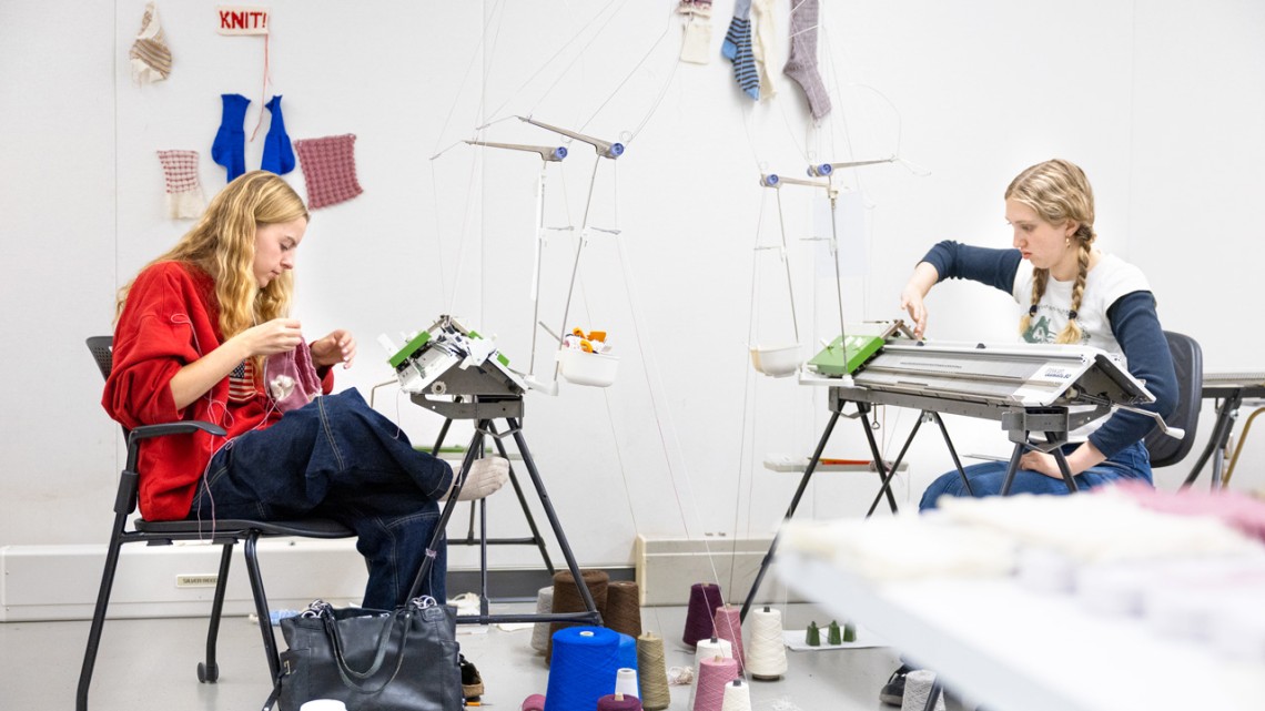 Fashion design majors Roan Harvey ’27, left, and Lucy Jones ’26 make knitted socks using wool grown and spun in New York state for the class Knitwear Design and Other Applications in the College of Human Ecology. Their design swatches and test socks are pinned to the wall.