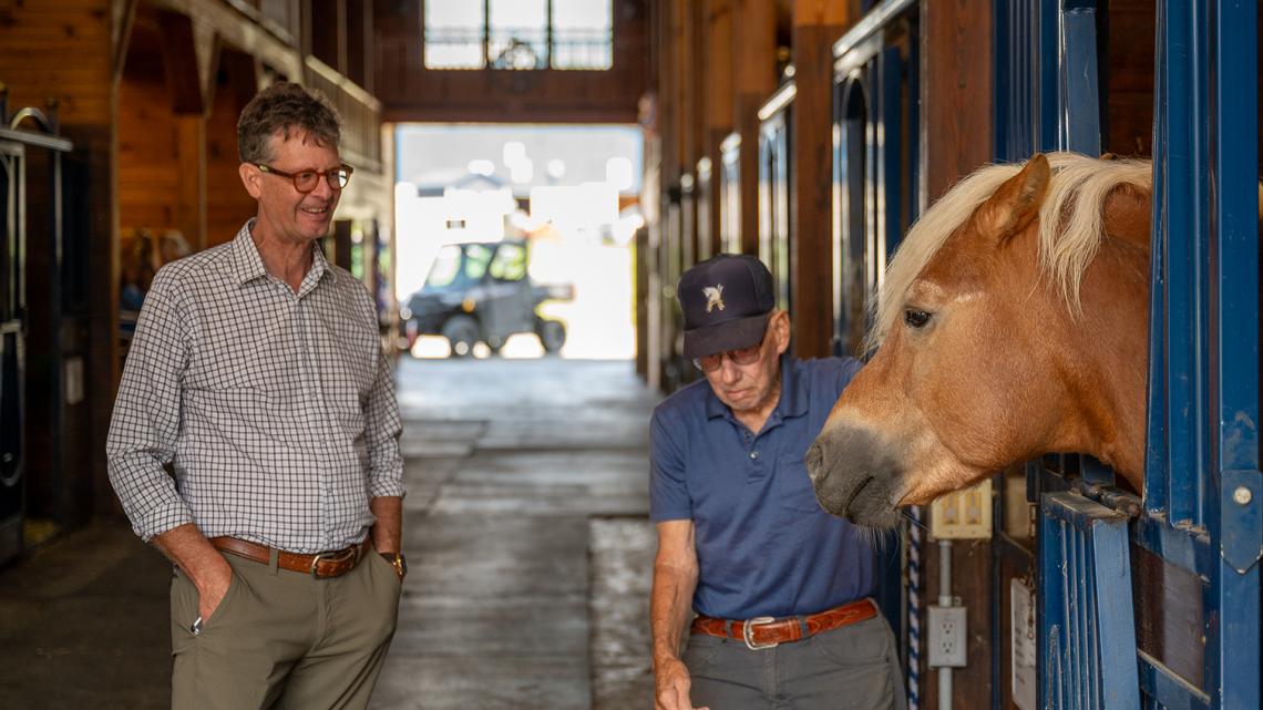 Two men stand in a sunlit horse stable beside a stall where a light brown horse with a blond mane leans its head out. One man, wearing glasses and a checked shirt, smiles with his hands in his pockets, while the other, in a navy cap and polo shirt, gestures toward the horse. The barn’s wooden walls and bright natural light create a warm, welcoming atmosphere.