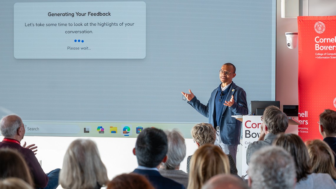 A man with short dark hair, glasses and a blue suit coat stands by a large screen in front of an audience.