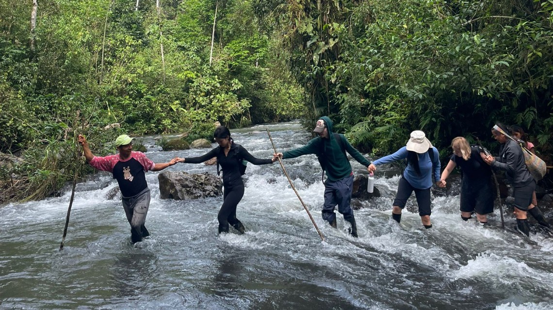 Laidlaw scholars hold hands while crossing a river. An Ecuadorian man leads the front of the line.
