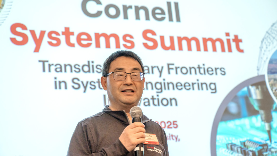 man with microphone speaks in front of sing that reads "Cornell systems summit"