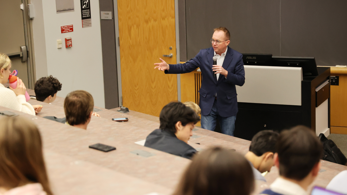 Director Mick Mulvaney stands at the front of a lecture hall speaking to Cornell students seated in rows,