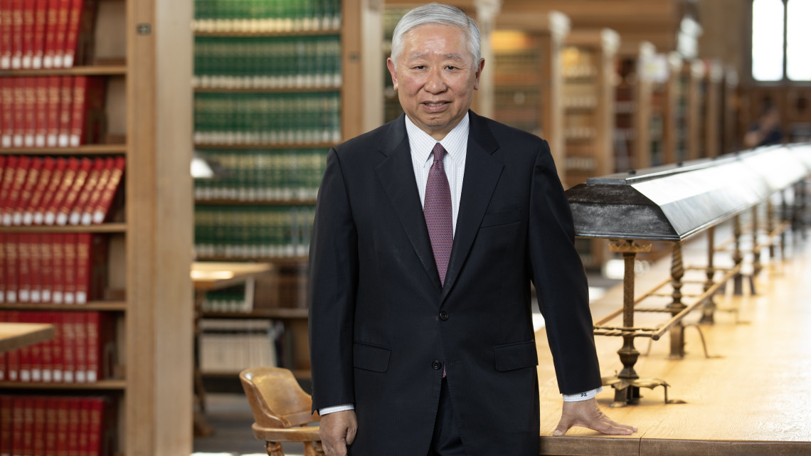 Jonathan Zhu stands in the Cornel Law Library with his hand on a long table and rows of shelves behind him