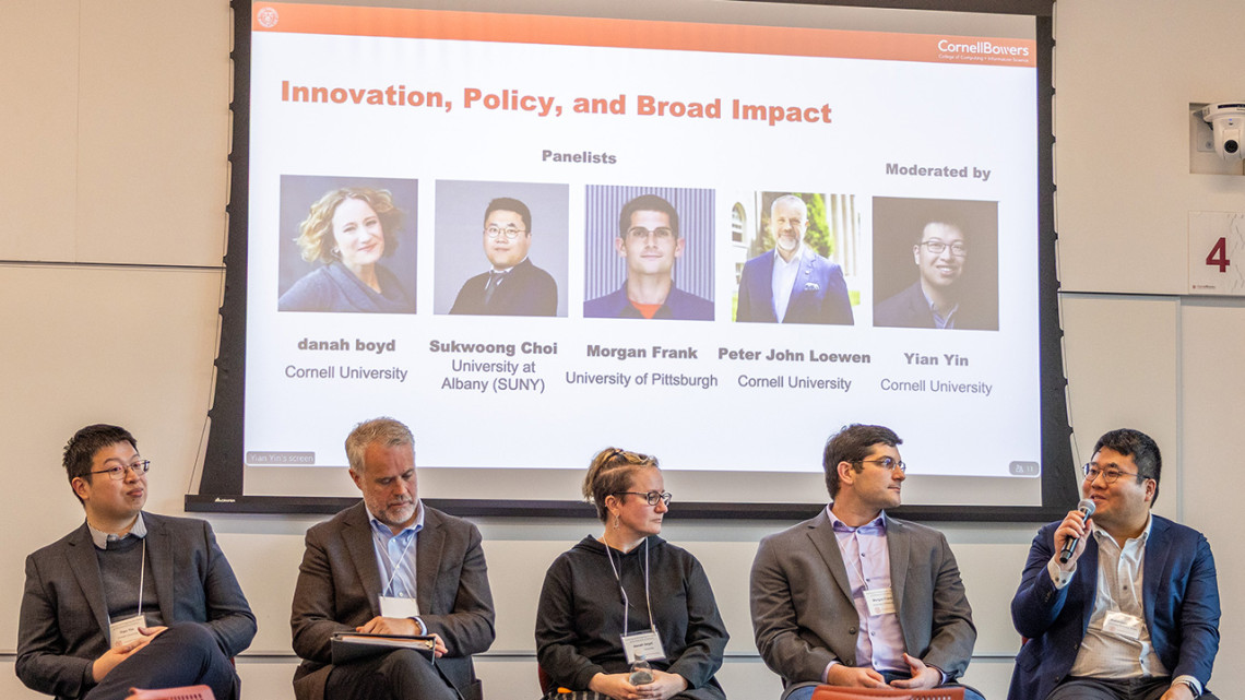 Five researchers sit in chairs in front of a screen as part of a panel discussion