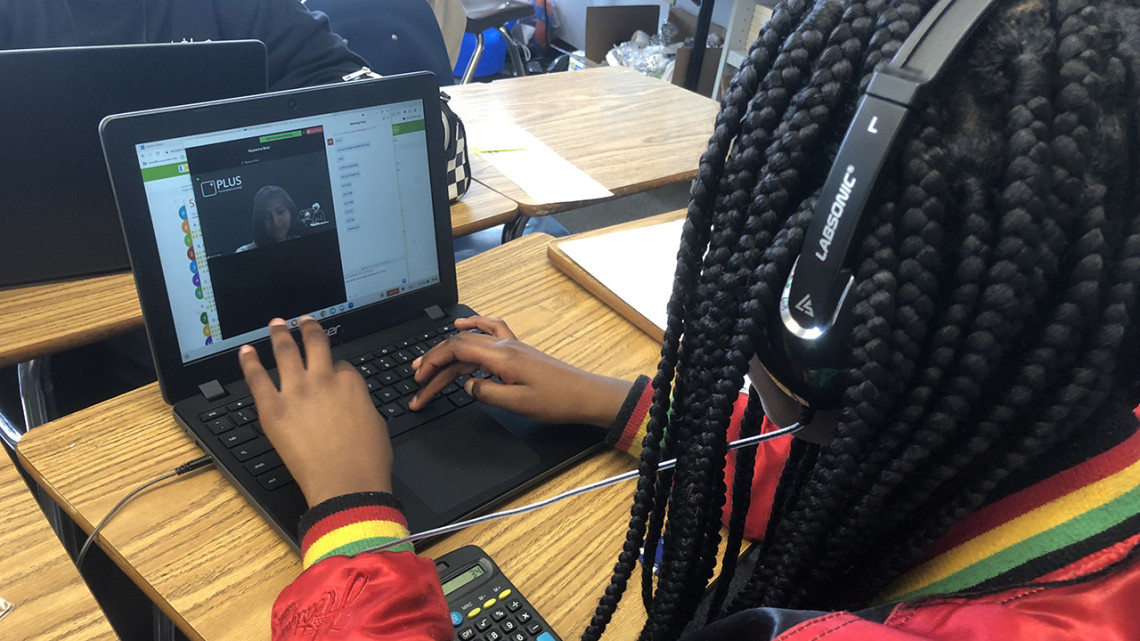 Seen from behind, a child with braids interacts with a tutor on a laptop screen