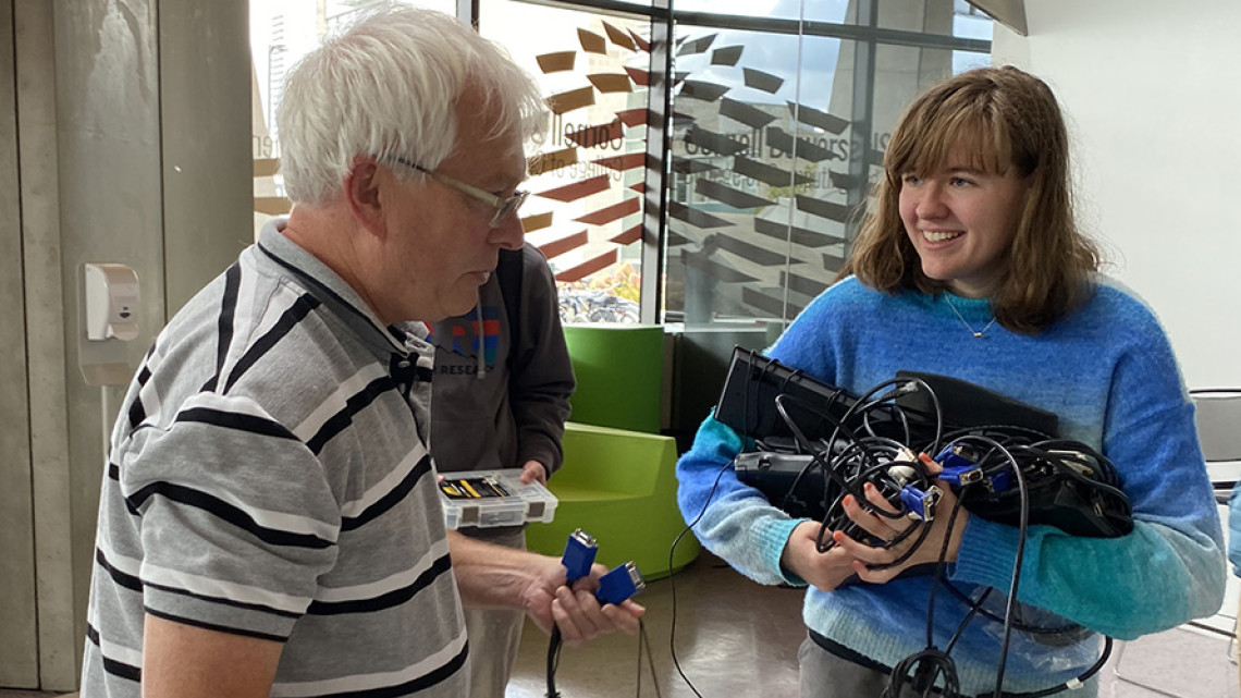 An older man talks to a youjg blonde woman i a blue sweater who us holding several cords and electronic devices.