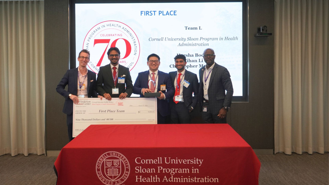 Harsha Boddu MHA ’27, Sloan MHA Faculty Director Professor Michael Richards, Tom Li MHA '27, Christopher Mathew MHA ’27 holding  a large check for first place  in front of a projector screen celebrating their win