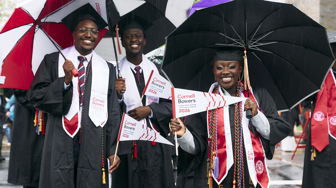 Three students stand in graduation regalia holding umbrellas and Cornell Bowers flags