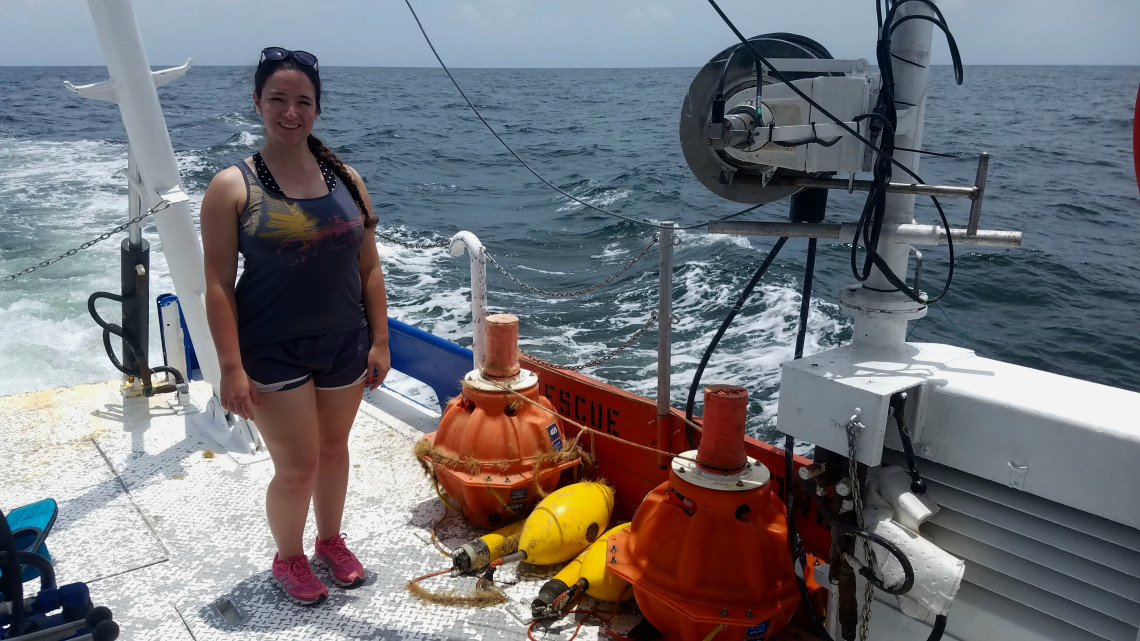 Dawn Parry stands on the deck of a research vessel in the Gulf of Mexico next to orange acoustic recording equipment recovered from the ocean and used to monitor whales and underwater noise.