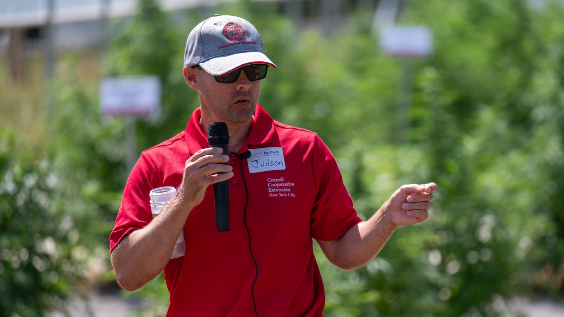Judson Reid speaks into a handheld microphone during an outdoor field day at Cornell AgriTech, gesturing toward research plots behind him.