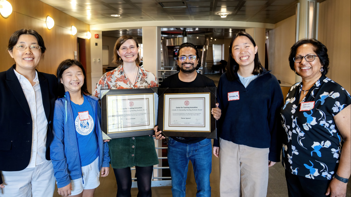 Six people pose indoors for a photo. The center two, a man and a woman, hold their award certificates for outstanding graduate teaching.