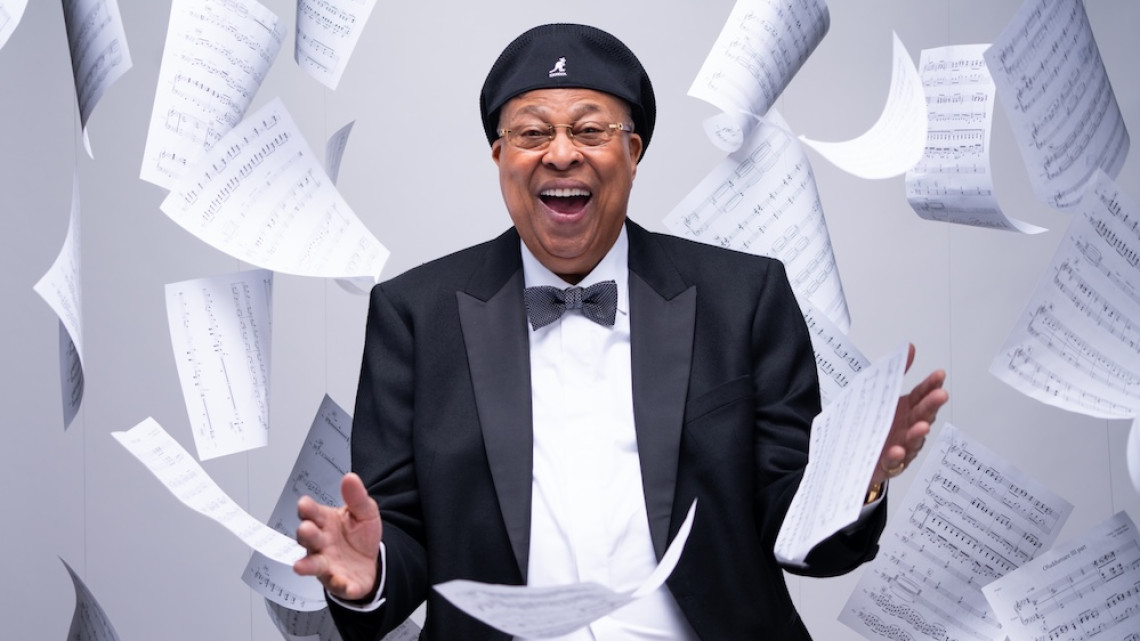 Person wearing a tuxedo and black cap smiles amidst a shower of sheet music